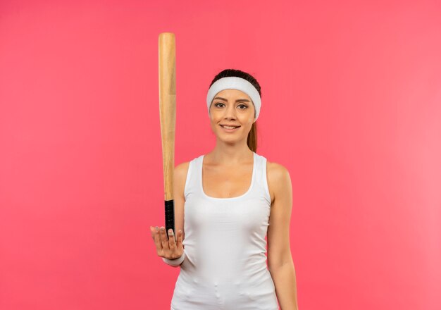 Jeune femme de remise en forme en tenue de sport avec bandeau tenant une chauve-souris avec sourire sur le visage debout sur un mur rose