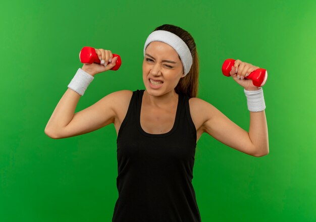 Jeune femme de remise en forme en tenue de sport avec bandeau tenant deux haltères, faire des exercices avec une expression agacée debout sur un mur vert