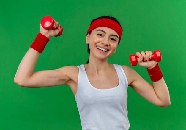 Jeune femme de remise en forme en tenue de sport avec bandeau tenant deux haltères levant les mains faisant des exercices souriant confiant debout sur mur vert