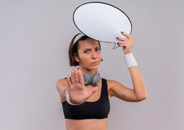 Jeune femme de remise en forme en tenue de sport avec bandeau tenant signe de bulle de discours vide faisant panneau d'arrêt avec la main avec un visage sérieux debout sur un mur blanc