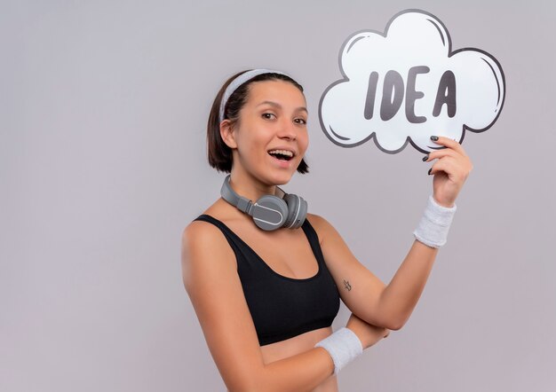 Jeune femme de remise en forme en tenue de sport avec bandeau tenant signe de bulle de parole avec idée de mot heureux et positif souriant joyeusement debout sur un mur blanc