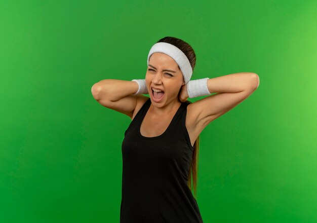Jeune femme de remise en forme en tenue de sport avec des cris avec une expression agacée de toucher son cou debout sur un mur vert