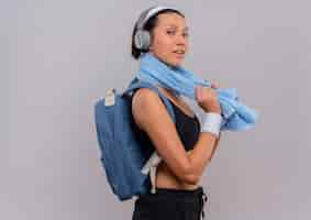 Photo gratuite jeune femme de remise en forme en tenue de sport avec sac à dos et casque sur la tête avec une serviette sur son cou à la confiance en souriant debout sur un mur blanc