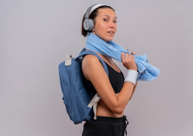 Jeune femme de remise en forme en tenue de sport avec sac à dos et casque sur la tête avec une serviette sur son cou à la confiance en souriant debout sur un mur blanc