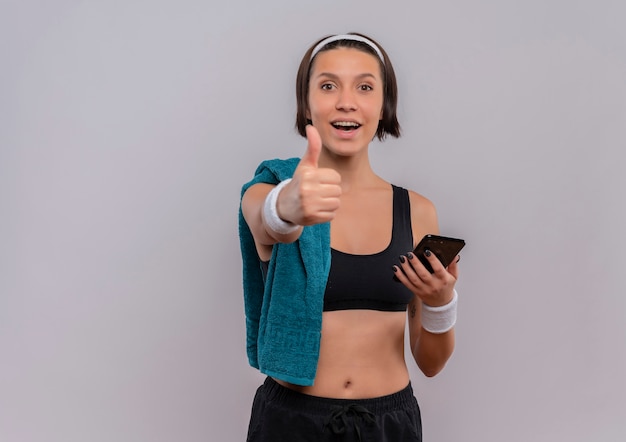 Jeune femme de remise en forme en tenue de sport avec une serviette sur l'épaule tenant le smartphone souriant montrant les pouces vers le haut debout sur un mur blanc