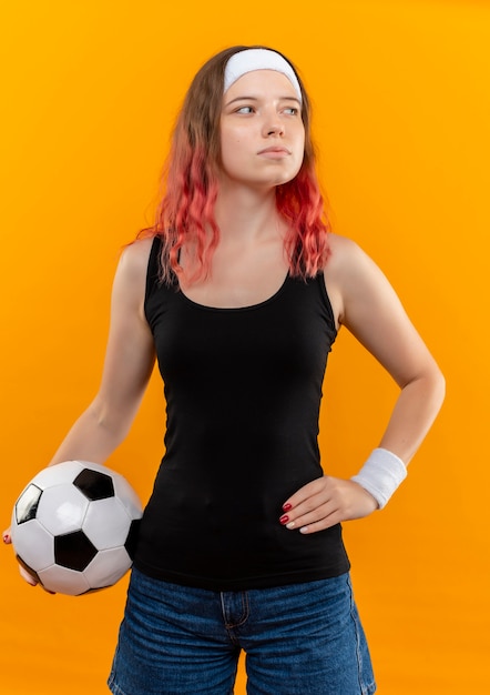 Photo gratuite jeune femme de remise en forme en tenue de sport tenant un ballon de soccer à côté avec un visage sérieux debout sur un mur orange