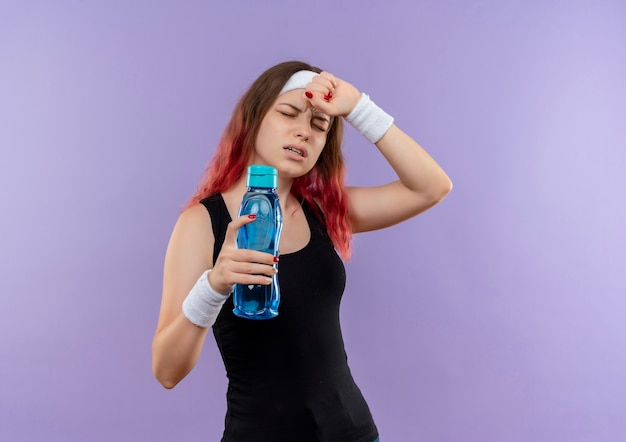 Jeune femme de remise en forme en tenue de sport tenant une bouteille d'eau à la recherche de mal avoir de forts maux de tête debout sur un mur violet