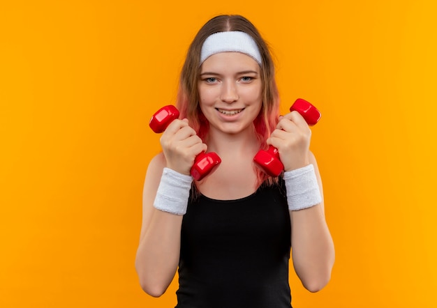 Jeune femme de remise en forme en tenue de sport tenant des haltères, faire des exercices souriant debout sur un mur orange