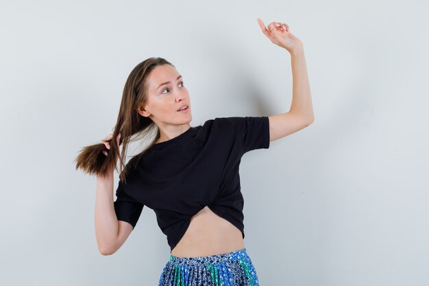 Jeune femme rentrant les cheveux et pointant vers le haut avec l'index, en détournant les yeux en t-shirt noir et jupe bleue et à la recherche concentrée