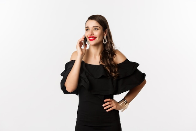 Jeune femme réussie en robe noire, rouge à lèvres et maquillage, parlant au téléphone portable et souriant, debout sur fond blanc