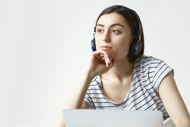 Jeune femme rêveuse dans des vêtements décontractés se détendre à la maison, écouter de la musique à l'aide de l'application en ligne sur ordinateur portable