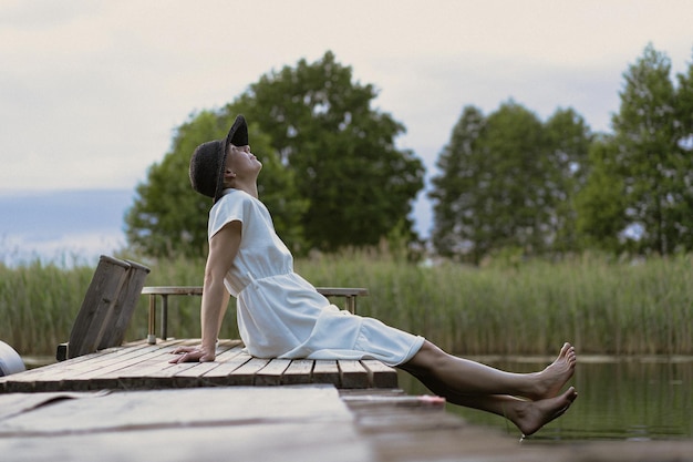 Jeune femme en robe et chapeau dans le parc au bord du lac à la jetée. Été, coucher de soleil, portrait