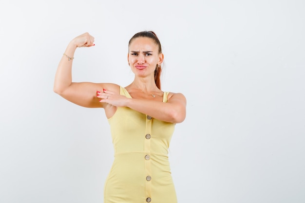 Photo gratuite jeune femme en robe jaune montrant les muscles du bras et l'air confiant, vue de face.