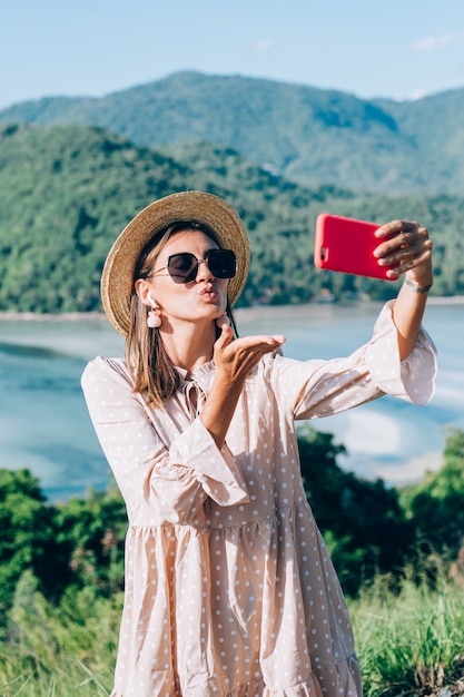 Jeune femme en robe mignonne d'été, chapeau de paille et lunettes de soleil faisant appel vidéo avec son smartphone