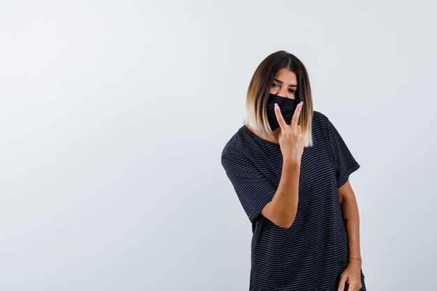 Jeune femme en robe noire, masque noir montrant le geste de paix et l'air confiant, vue de face.