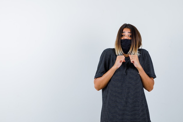 Photo gratuite jeune femme en robe noire, masque noir serrant les poings et à la peur, vue de face.