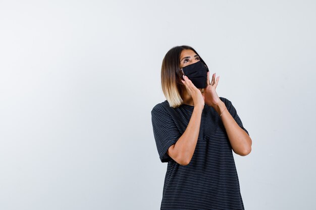 Jeune femme en robe noire, masque noir tenant par la main près de la bouche pour appeler quelqu'un et à la recherche focalisée, vue de face.