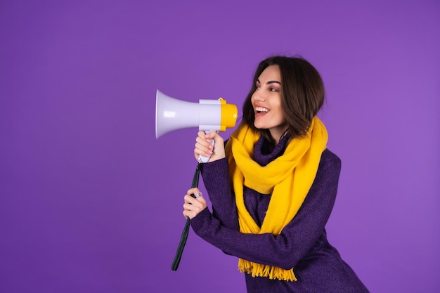 Photo gratuite jeune femme en robe tricotée et écharpe jaune sur fond violet crie joyeusement dans un mégaphone, joyeuse, annonce une vente