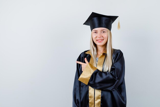 Jeune femme en robe universitaire pointant de côté et ayant l'air heureuse