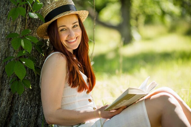 Jeune femme romantique au chapeau de paille lisant un livre assis dans le jardin