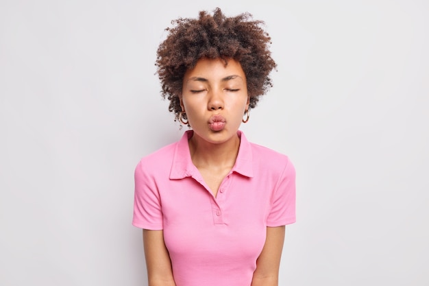 Une jeune femme romantique aux cheveux bouclés se penche en avant ferme les yeux et les lèvres plissées attendent le baiser anticipe que mwah exprime des sentiments véridiques porte un t-shirt rose décontracté isolé sur un mur blanc