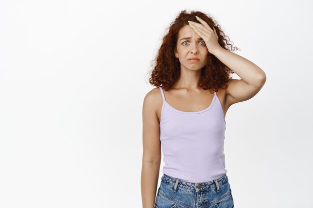 Jeune femme rousse bouleversée tient la main sur le front, a du mal, a oublié de se souvenir de qch, l'air affligé, choqué et sombre, debout sur fond blanc