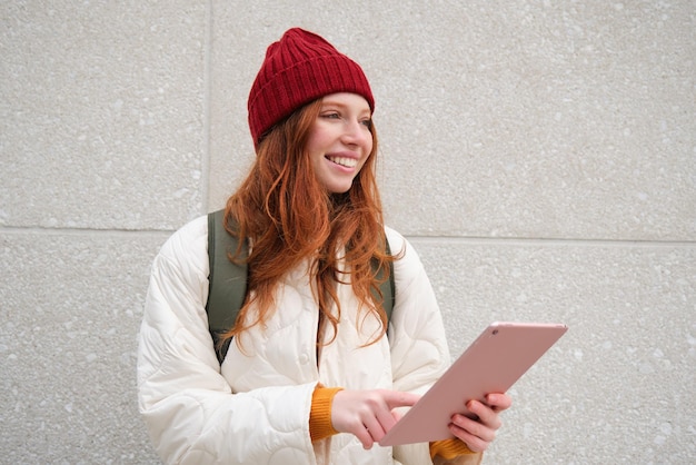 Jeune femme rousse avec un chapeau rouge utilise sa tablette numérique à l'extérieur se dresse sur la rue avec gadget conne