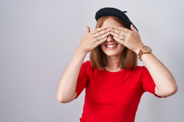 Photo gratuite jeune femme rousse debout portant des lunettes et un béret couvrant les yeux avec les mains souriant concept aveugle joyeux et drôle