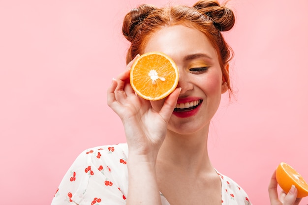 Jeune femme rousse énergique en T-shirt blanc tenant de savoureuses oranges sur fond rose.