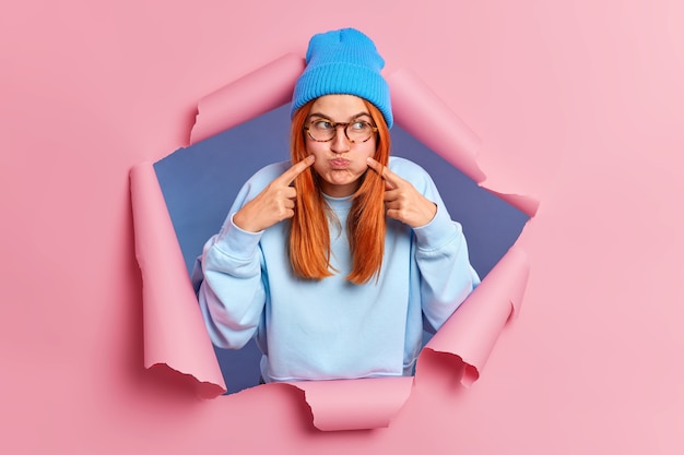 Photo gratuite la jeune femme rousse fait la grimace moue les joues retient les points de souffle des index sur le visage des fous autour porte des lunettes chapeau bleu et un pull.