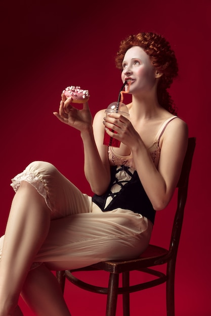 Photo gratuite jeune femme rousse médiévale comme une duchesse en corset noir et vêtements de nuit assis sur une chaise sur l'espace rouge avec un verre et un beignet