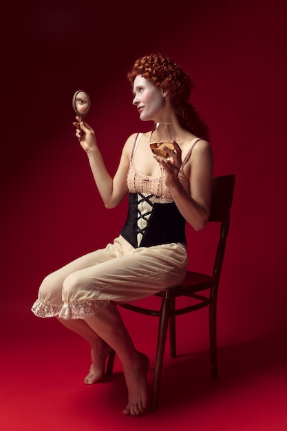Photo gratuite jeune femme rousse médiévale en tant que duchesse en corset noir et vêtements de nuit assis sur un espace rouge avec un miroir et un verre de vin