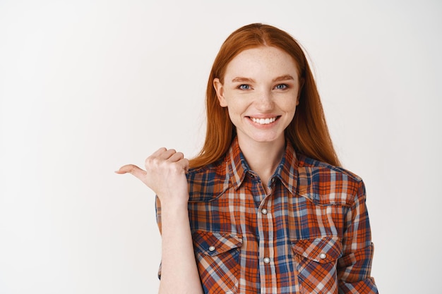 Jeune femme rousse pointant le pouce vers la gauche et souriante, montrant une offre promotionnelle, mur blanc