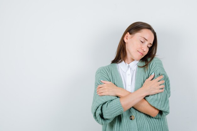 Jeune femme s'embrassant en chemise, cardigan et semblant paisible, vue de face.