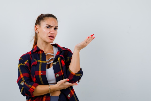 Jeune femme s'étirant la main dans un geste de questionnement en haut court, chemise à carreaux et semblant anxieuse. vue de face.