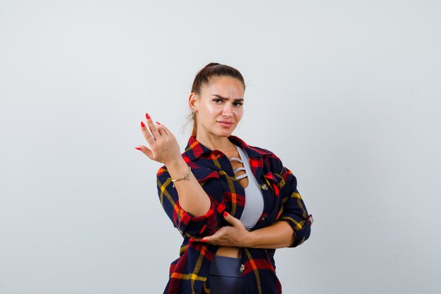 Jeune femme s'étirant la main dans un geste de questionnement en haut court, chemise à carreaux et semblant sérieuse, vue de face.