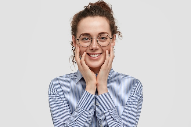 Une jeune femme satisfaite positive a un sourire doux, touche les joues avec les mains