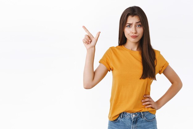 Une jeune femme sceptique, arrogante et mécontente trouve un nouveau produit suspect et indigne de son attention, sourit avec dédain, fronçant les sourcils, jugeant, pointant vers le coin supérieur gauche Espace de copie