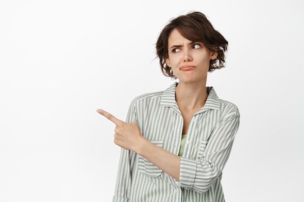 Jeune femme sceptique et suspecte pointant et regardant à gauche avec un sourcil levé, incertaine, ayant des doutes, debout sur fond blanc.