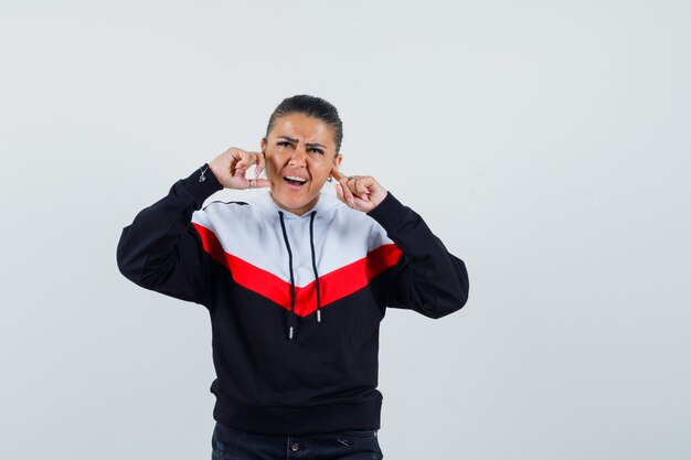 Jeune femme se bouchant les oreilles avec les doigts en sweat-shirt coloré et à l'ennui. vue de face.