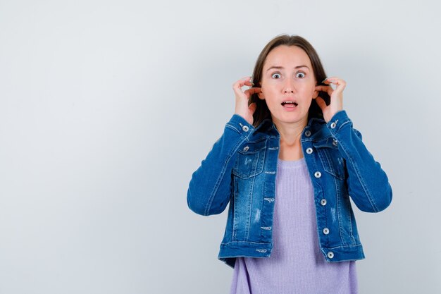 Jeune femme se branchant les oreilles avec les doigts en t-shirt, veste et à la perplexité. vue de face.