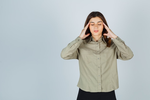 Jeune femme se frottant les tempes en chemise, jupe et l'air fatigué, vue de face.