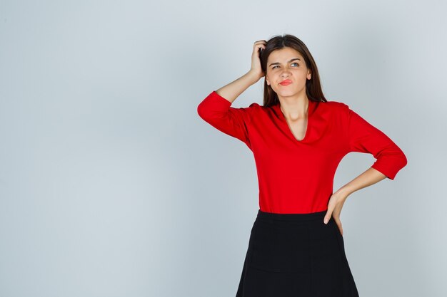 Jeune femme se gratter la tête tout en gardant la main sur la hanche en chemisier rouge