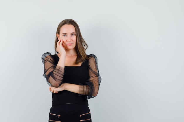 Jeune femme se penchant les joues sur la paume et posant à la caméra en chemisier noir et pantalon noir et à la charmante. vue de face.