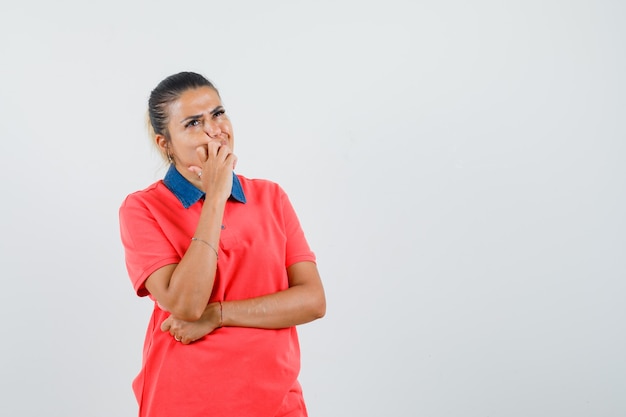 Jeune femme se penchant la main sur la bouche, debout dans la pensée pose en t-shirt rouge et à la pensif. vue de face.