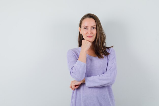 Jeune femme se penchant sur son poing tout en souriant à la caméra en chemisier lilas et à la fraîcheur