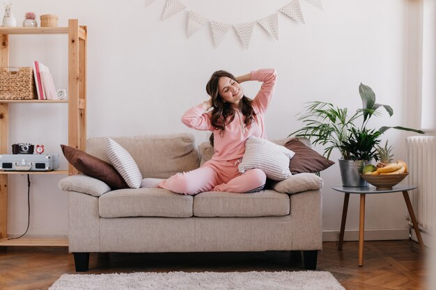 Jeune femme se repose dans sa chambre, assise sur un canapé, entouré d'étagères et d'une table avec pot de fleur