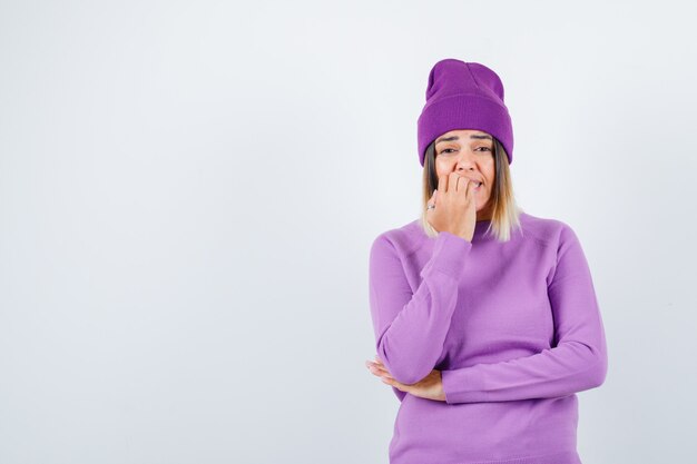 Jeune femme se rongeant les ongles en pull violet, bonnet et à l'air excité, vue de face.