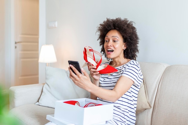 Jeune femme se sentant excitée par de nouvelles chaussures déballant ses achats et prenant des photos avec son téléphone à la maison en les publiant sur les réseaux sociaux