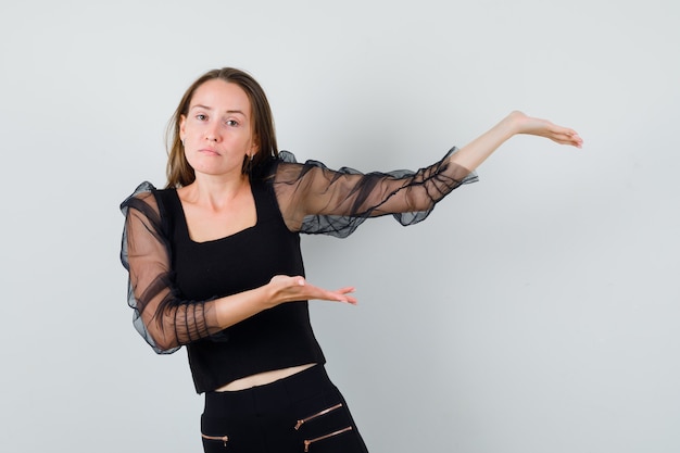 Jeune femme se tenant la main comme présentant quelque chose en chemisier noir et pantalon noir et à la sérieuse